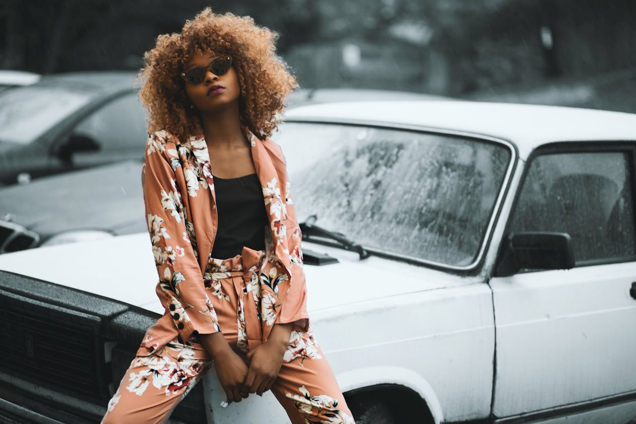 Fashionable woman in floral suit poses confidently on a classic car, showcasing vintage style.
